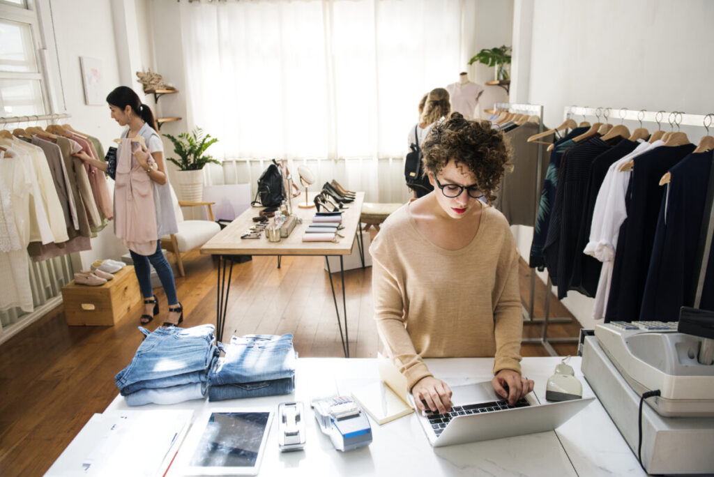 mulher mexendo no computador com funcionarias ao fundo mexendo em roupas numa arara - digital commerce